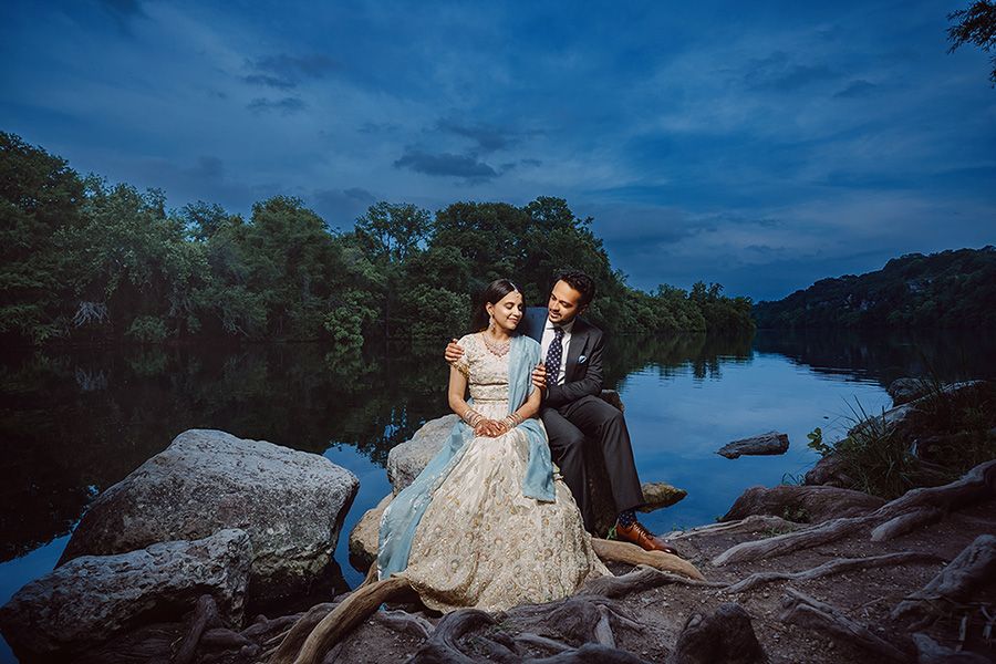 Couple in elegant wedding attire sitting on rocks by a serene river at dusk with lush trees and dramatic blue sky.