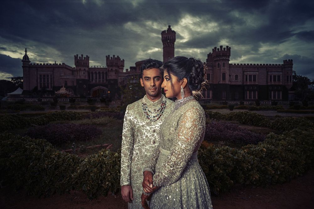 Couple in ornate traditional Indian attire standing before a castle at dusk.