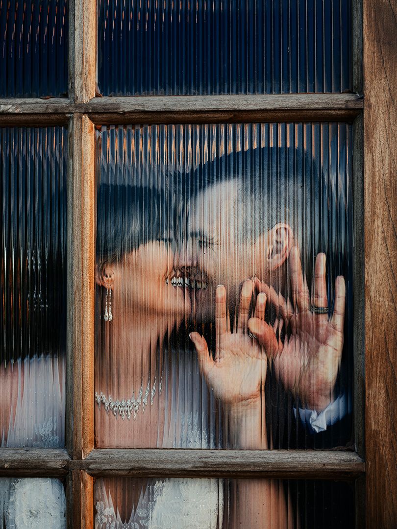 A couple shares a romantic kiss behind a textured glass window, hands pressed against the pane, wearing wedding attire.
