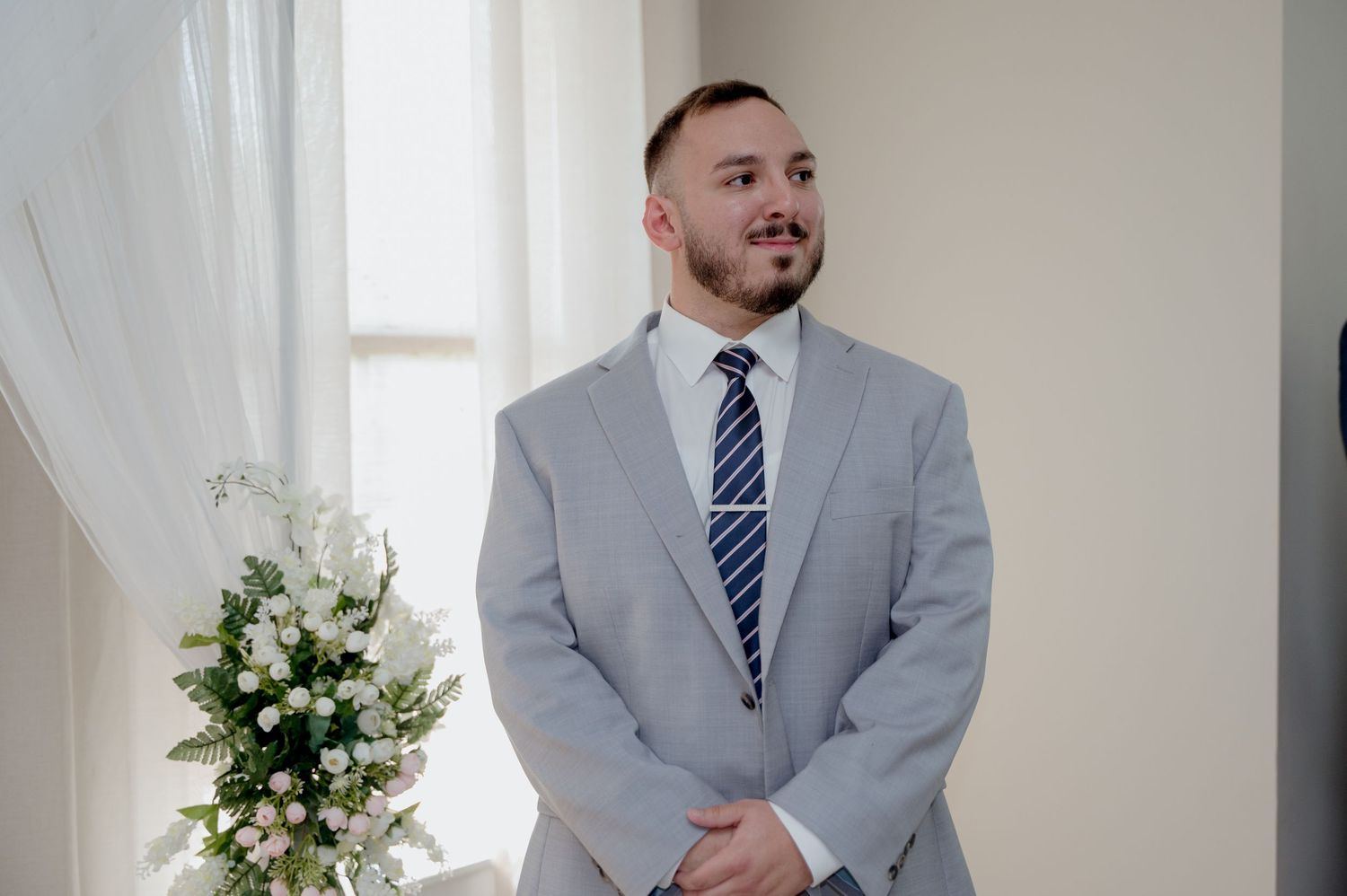 A person in a grey suit and blue striped tie poses during a formal photo session at a venue with white curtains.