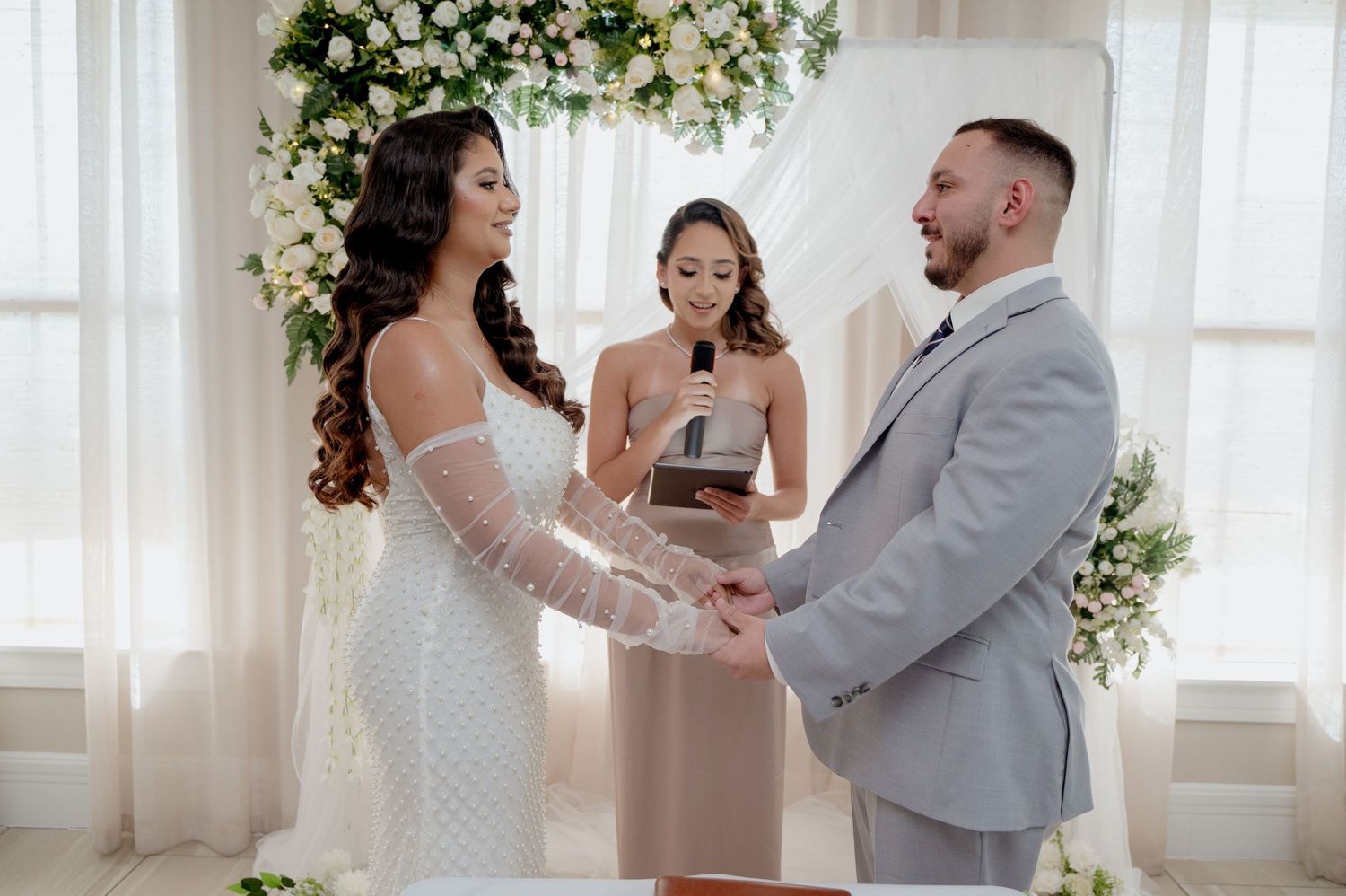 A couple exchanges vows during an indoor wedding ceremony beneath a romantic floral arch with white draping.