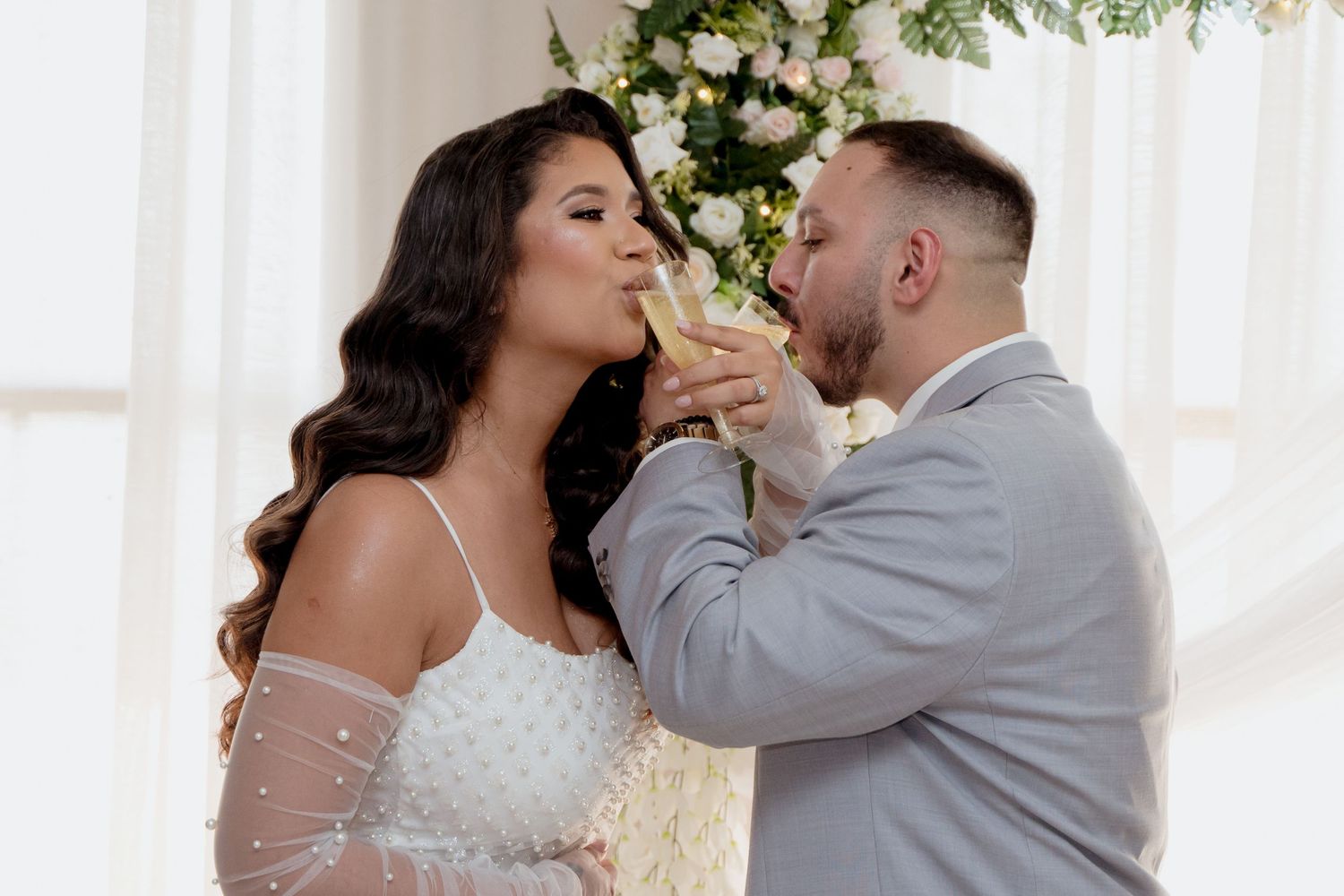 Affectionate bride and groom embrace under white floral arch decorations during indoor wedding ceremony.