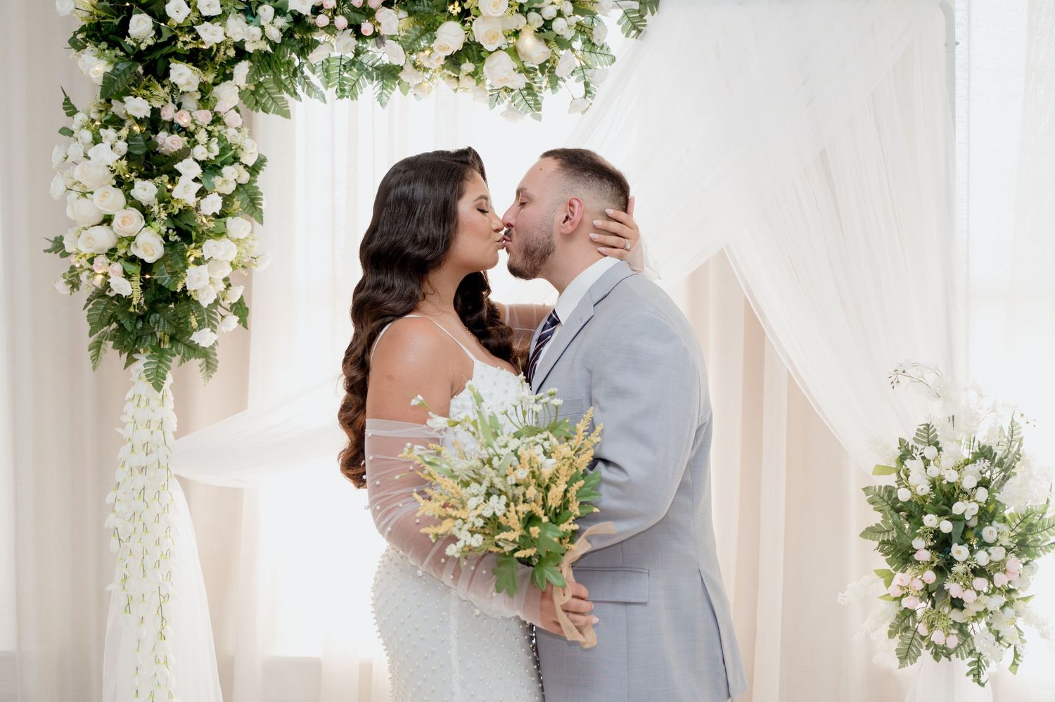 A romantic kiss under a white floral arch during a spring wedding ceremony with elegant white and green decor.