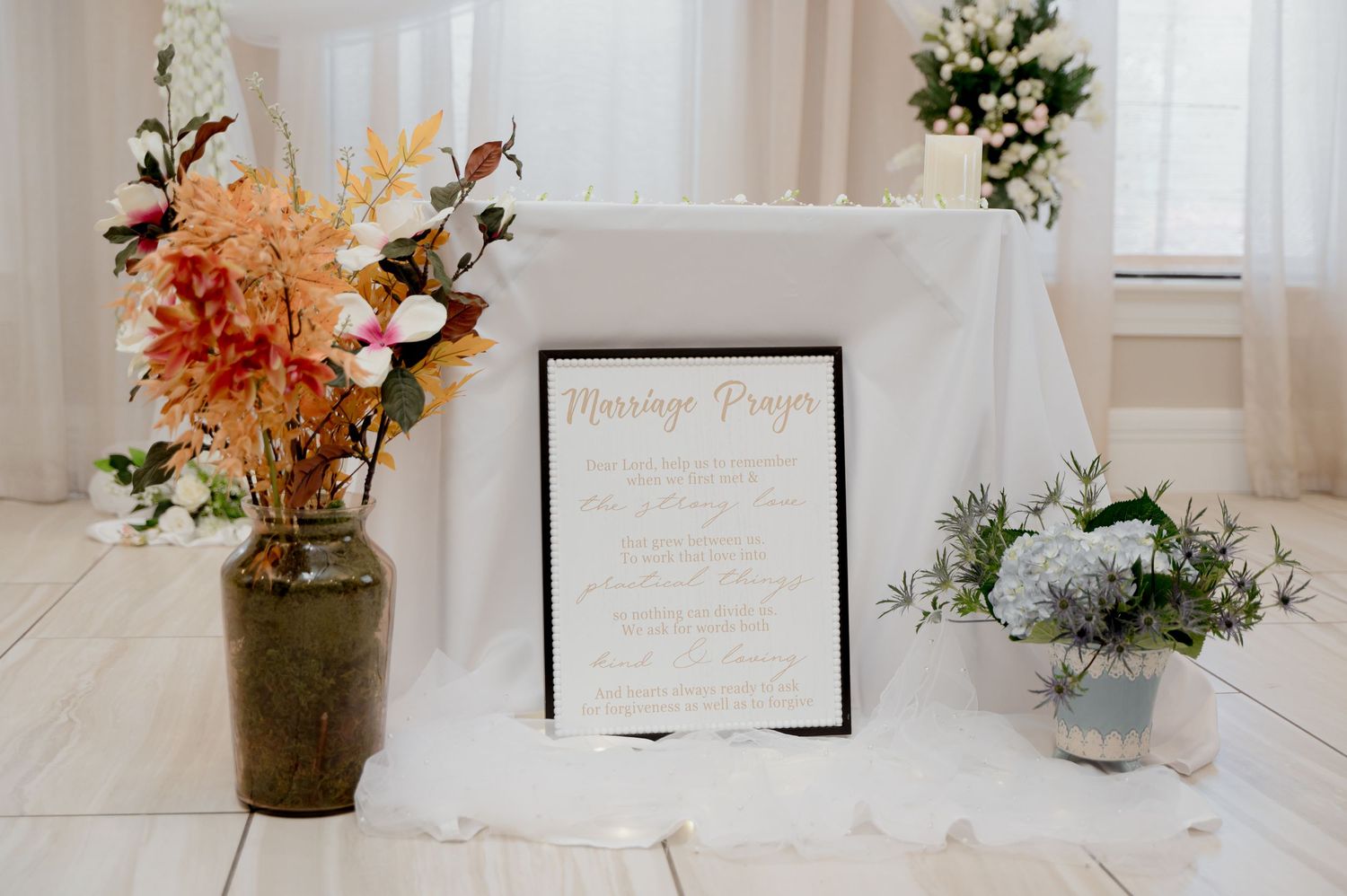 Elegant wedding table setup with framed menu card and floral arrangements in rustic vases against white curtains.