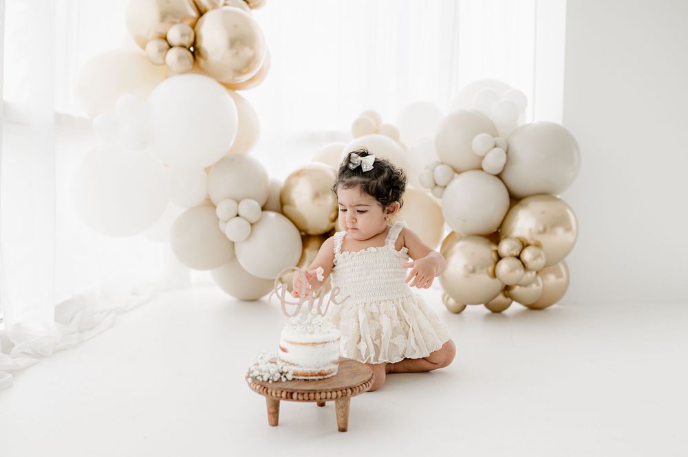 Baby photoshoot in white studio with decorative balloon arch and wooden stool backdrop.