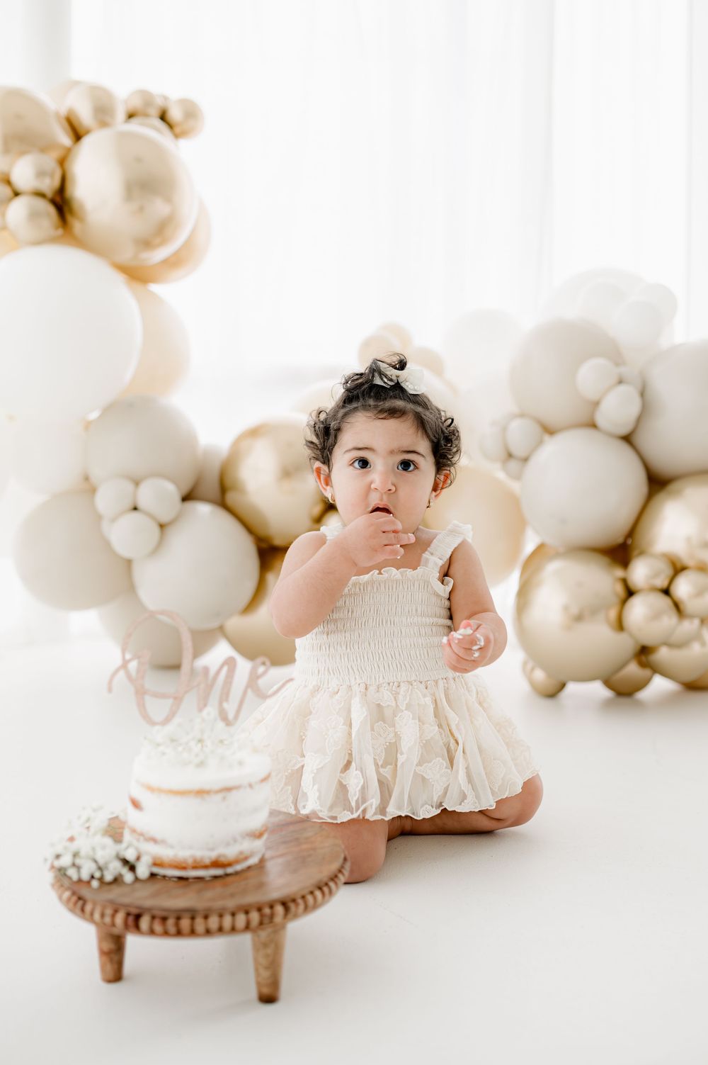 Baby in white lace dress sits near birthday cake with elegant gold and white balloon arch backdrop.