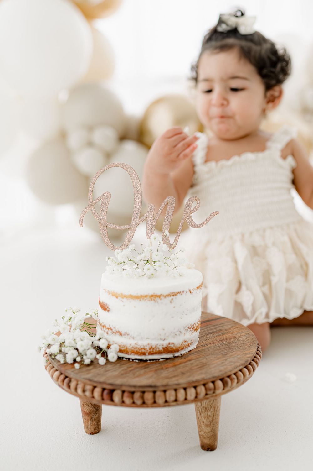 A white frosted cake with baby's breath flowers sits atop a wooden pedestal stand in a bright white setting.