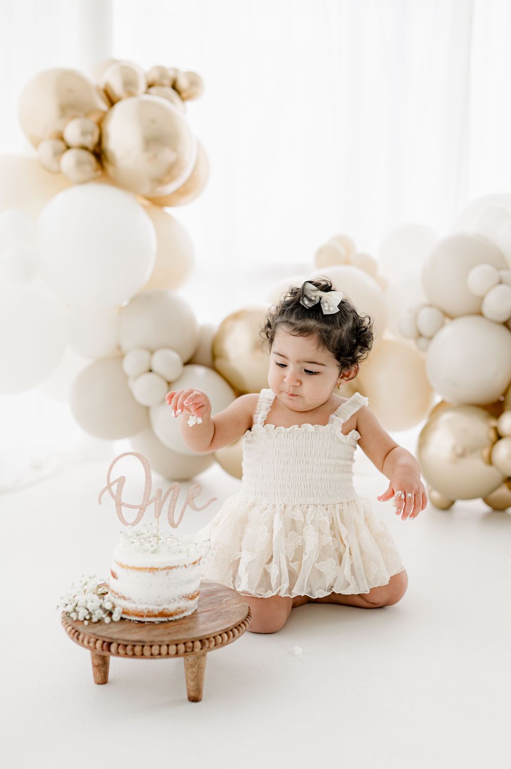 First birthday celebration with white dress, neutral balloon arch decor and small 'One' cake on wooden stand.