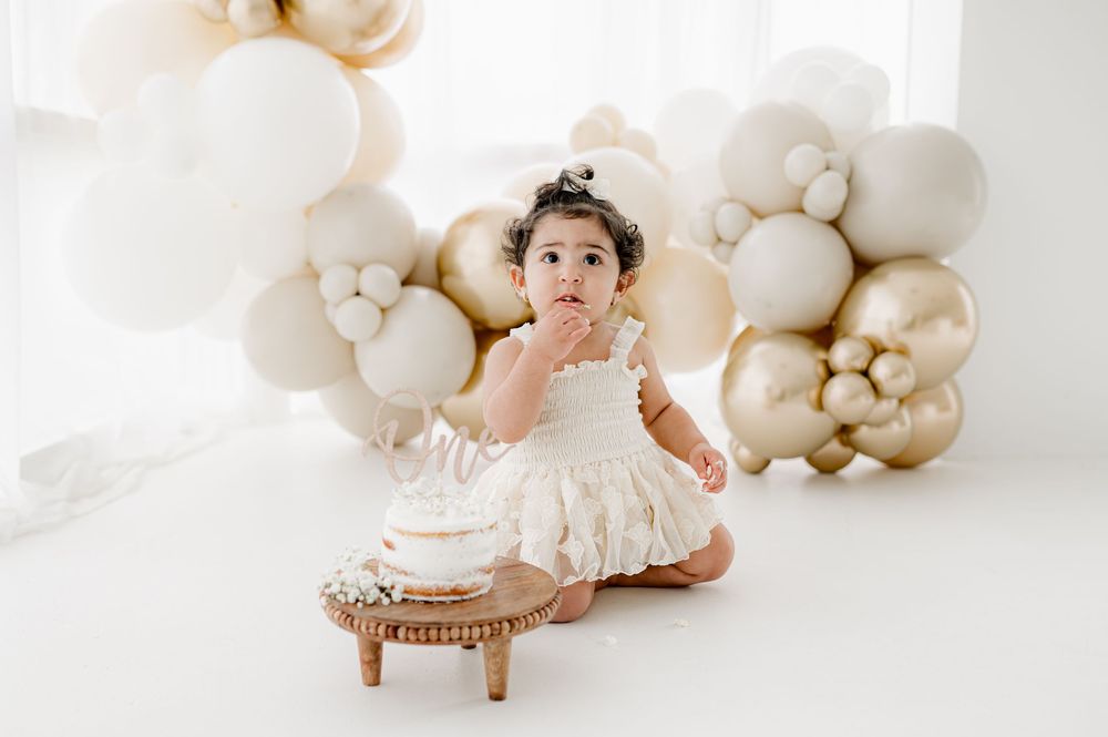 A baby in a white dress sits on a wooden stool against a backdrop of elegant beige and white balloon decorations.