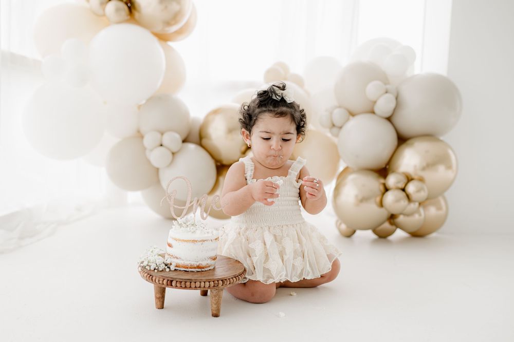 A small child in a white lace dress sits by a cake with elegant white and gold balloon decorations in the background.