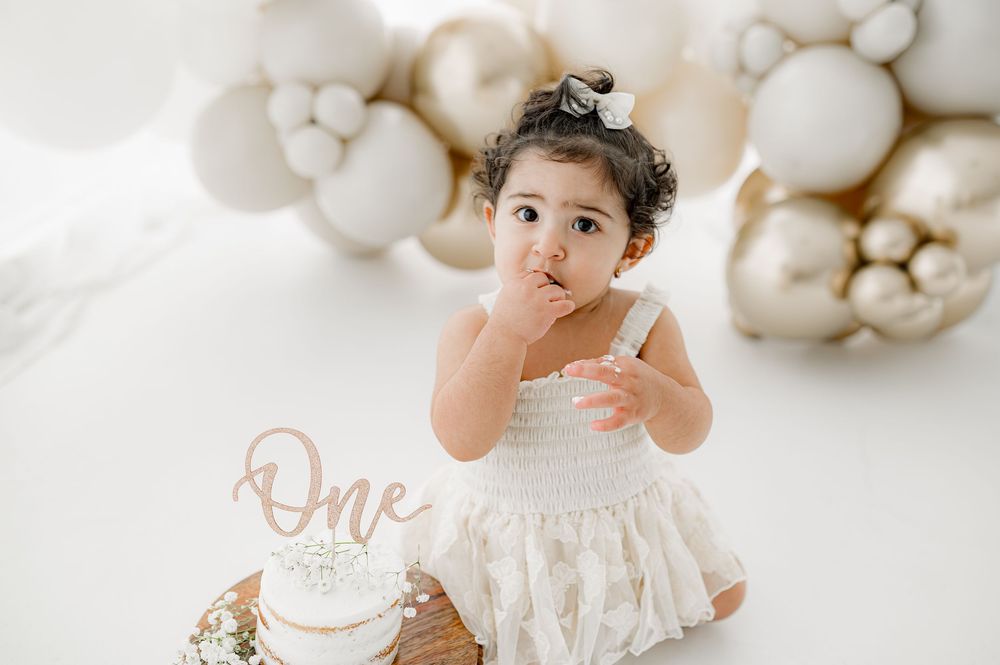 Baby in white lace dress poses with white balloons and birthday cake during elegant cake smash photo session.