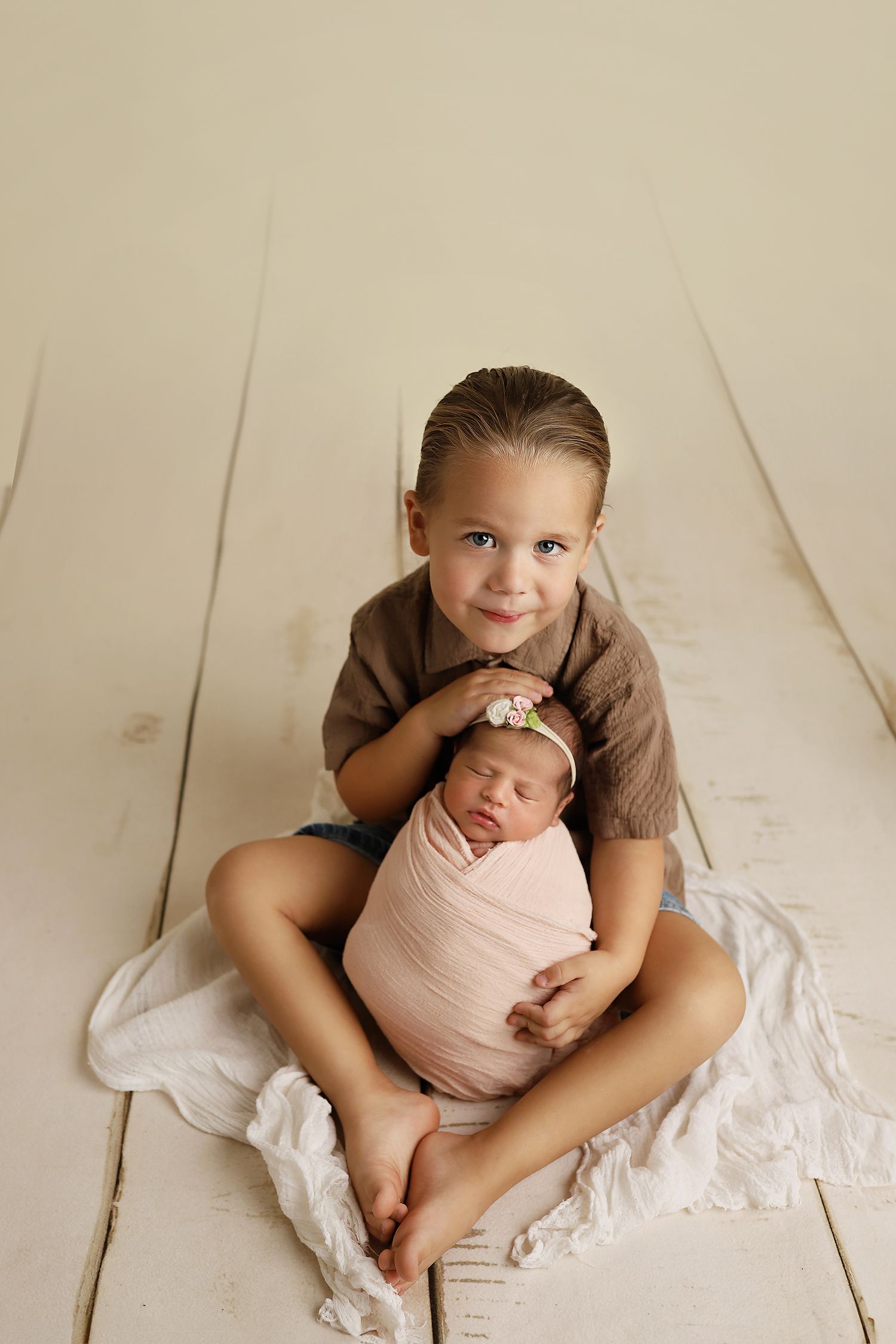 Child sitting cross-legged on wooden floor holding sleeping swaddled newborn in pink wrap against cream backdrop.