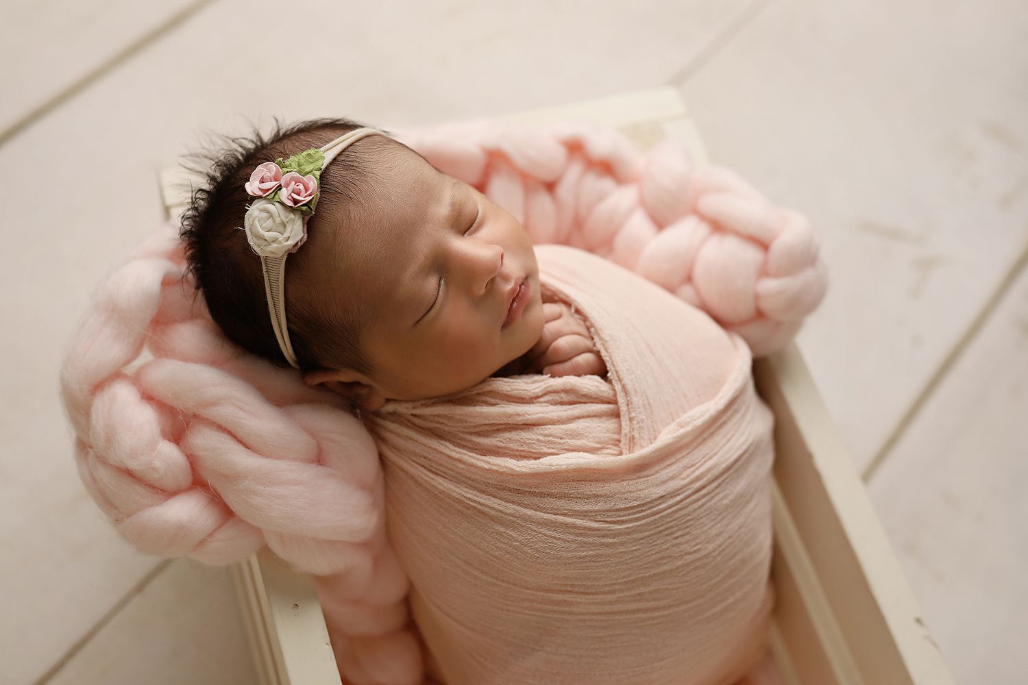 Sleeping newborn baby wrapped in peach fabric with floral headband rests on light wooden floor.