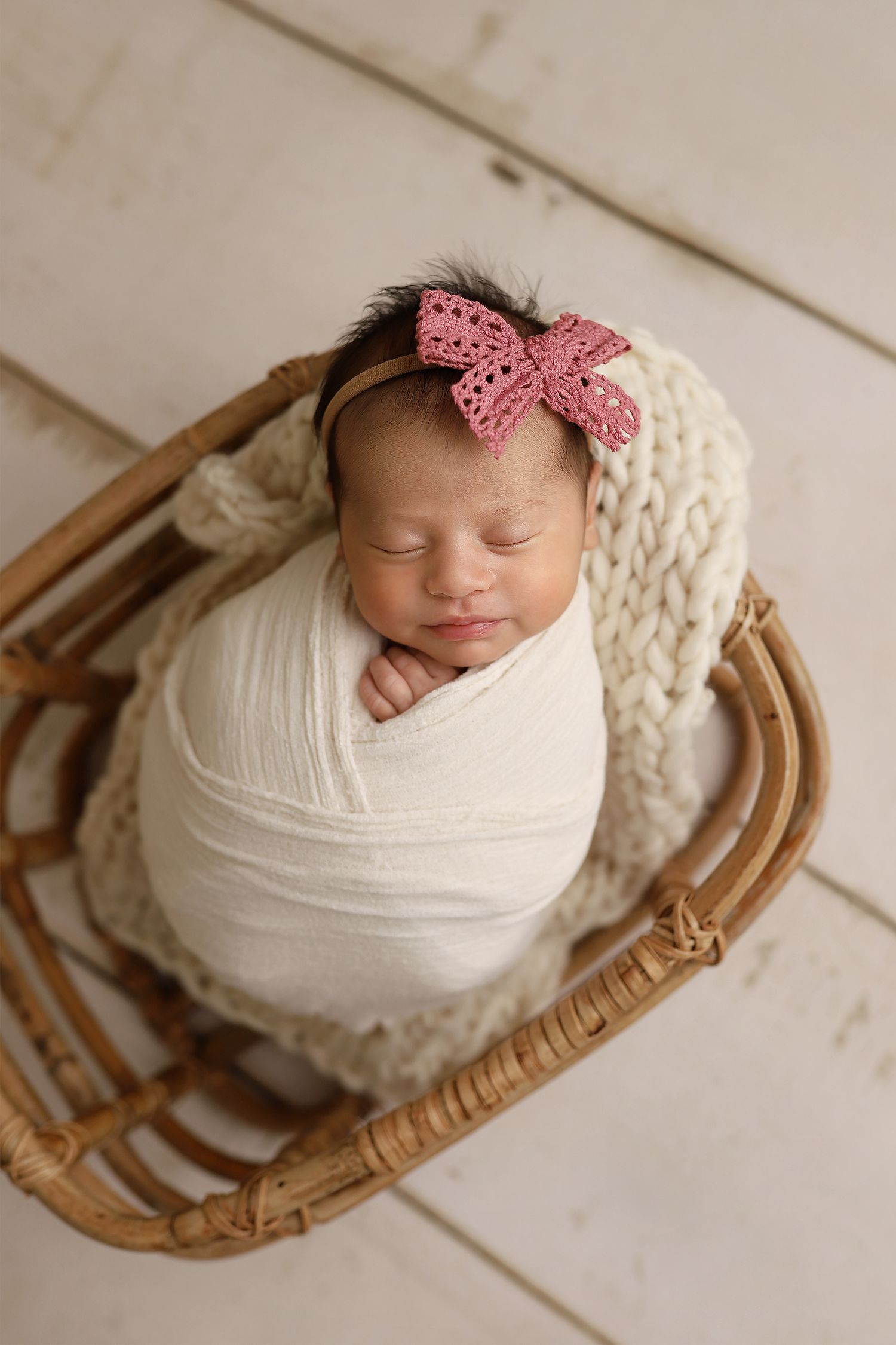 A tiny newborn baby wrapped in soft white cloth sleeps peacefully in a wooden basket with a pink hair bow. Utah newborn photographer