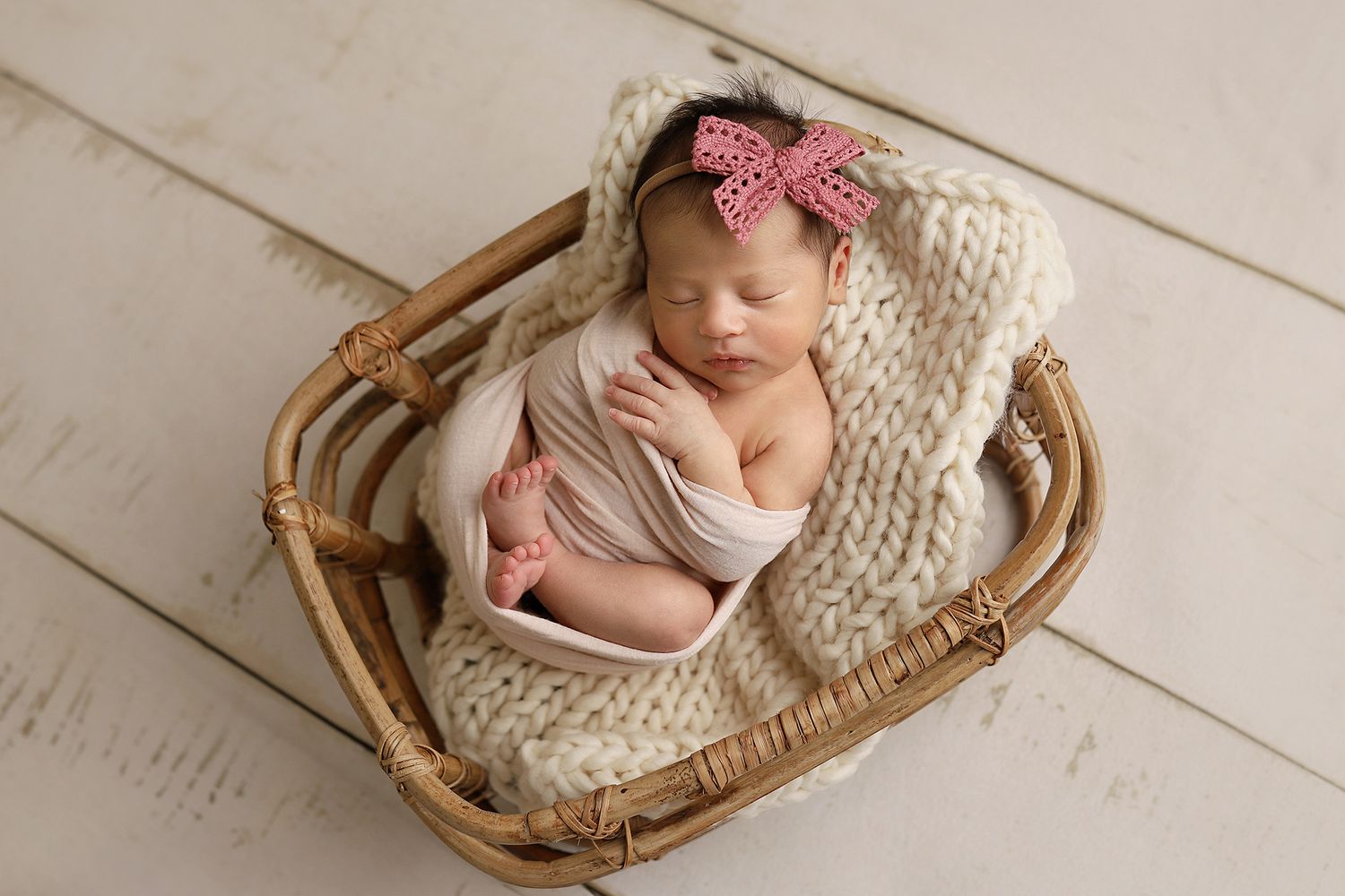 Sleeping newborn wrapped in soft fabric rests peacefully in wicker basket with knit blanket and pink bow. Orem newborn photographer