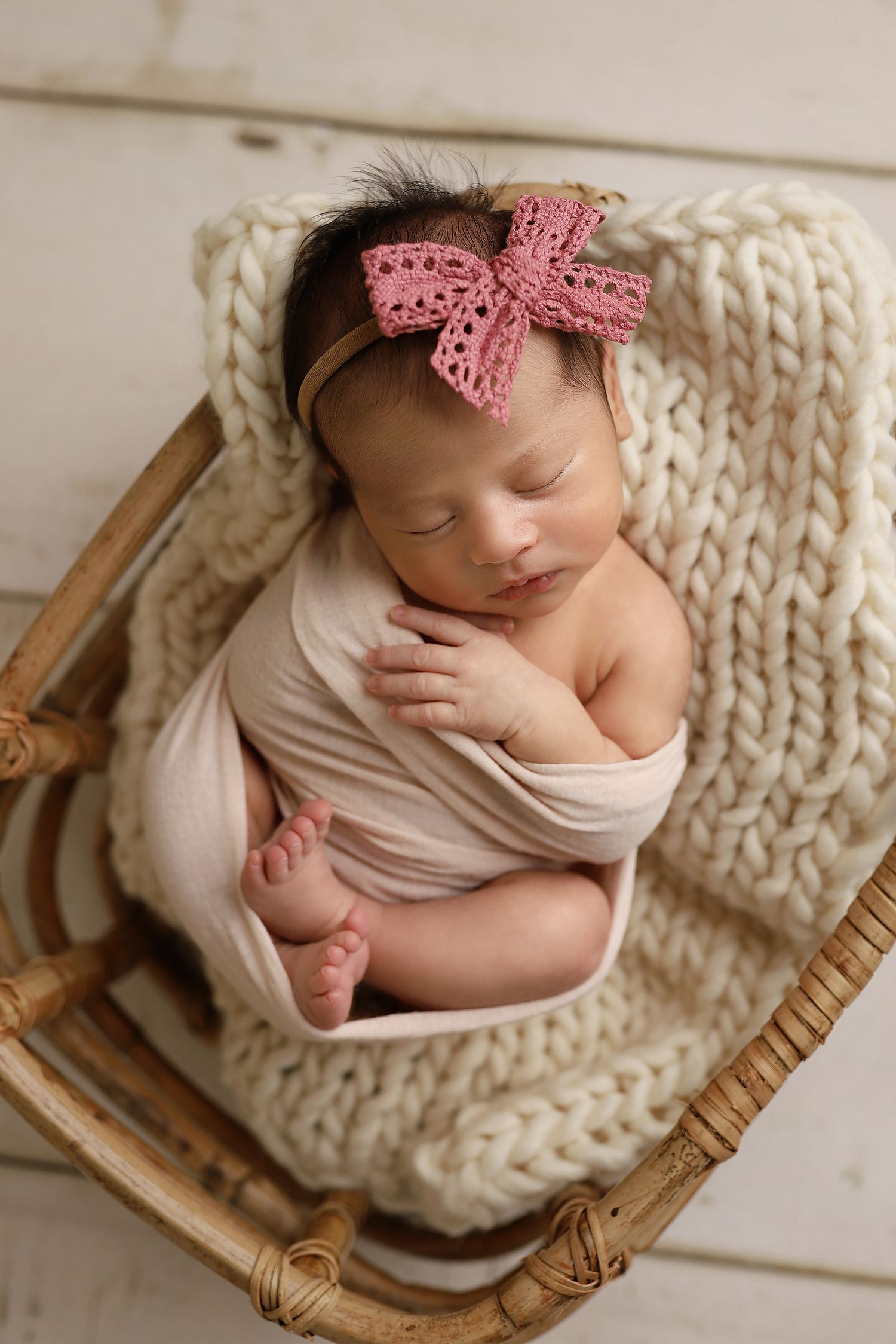 Newborn baby sleeping peacefully in wicker basket with chunky knit blanket and pink bow headband. Utah newborn photographer