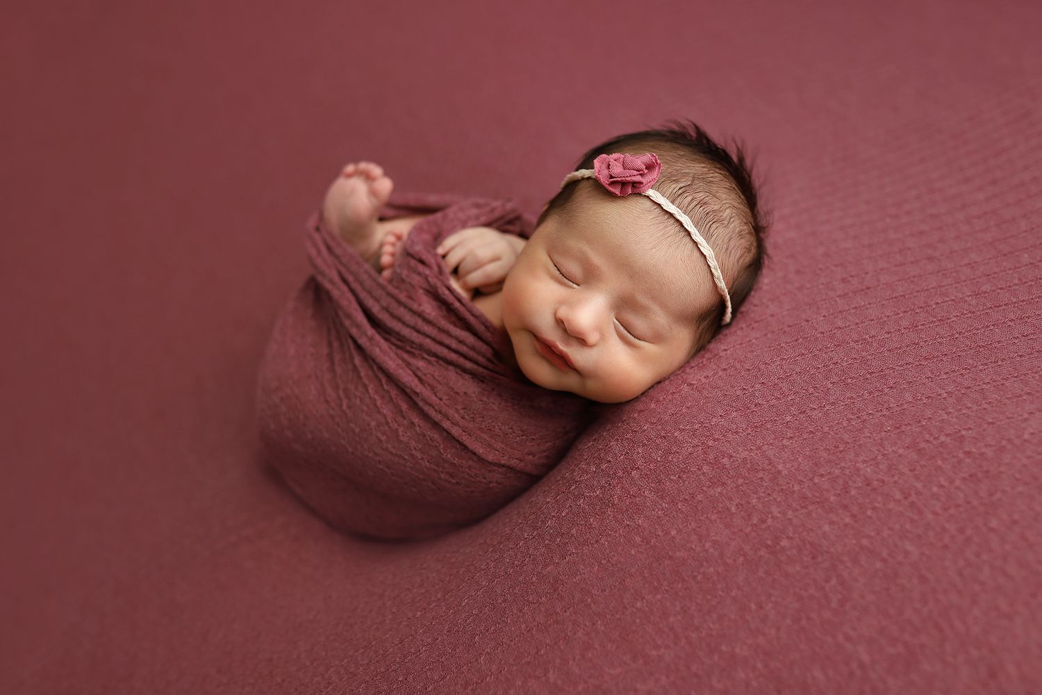 A newborn baby nestled in soft burgundy fabric during a professional photo session.