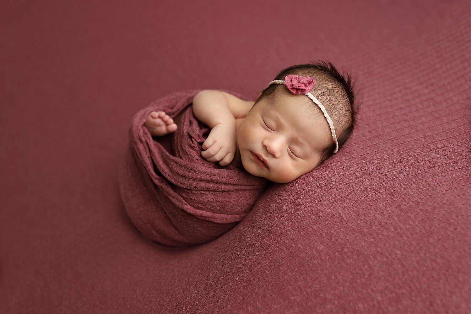 Newborn baby wrapped in burgundy blanket sleeping peacefully on matching burgundy backdrop.