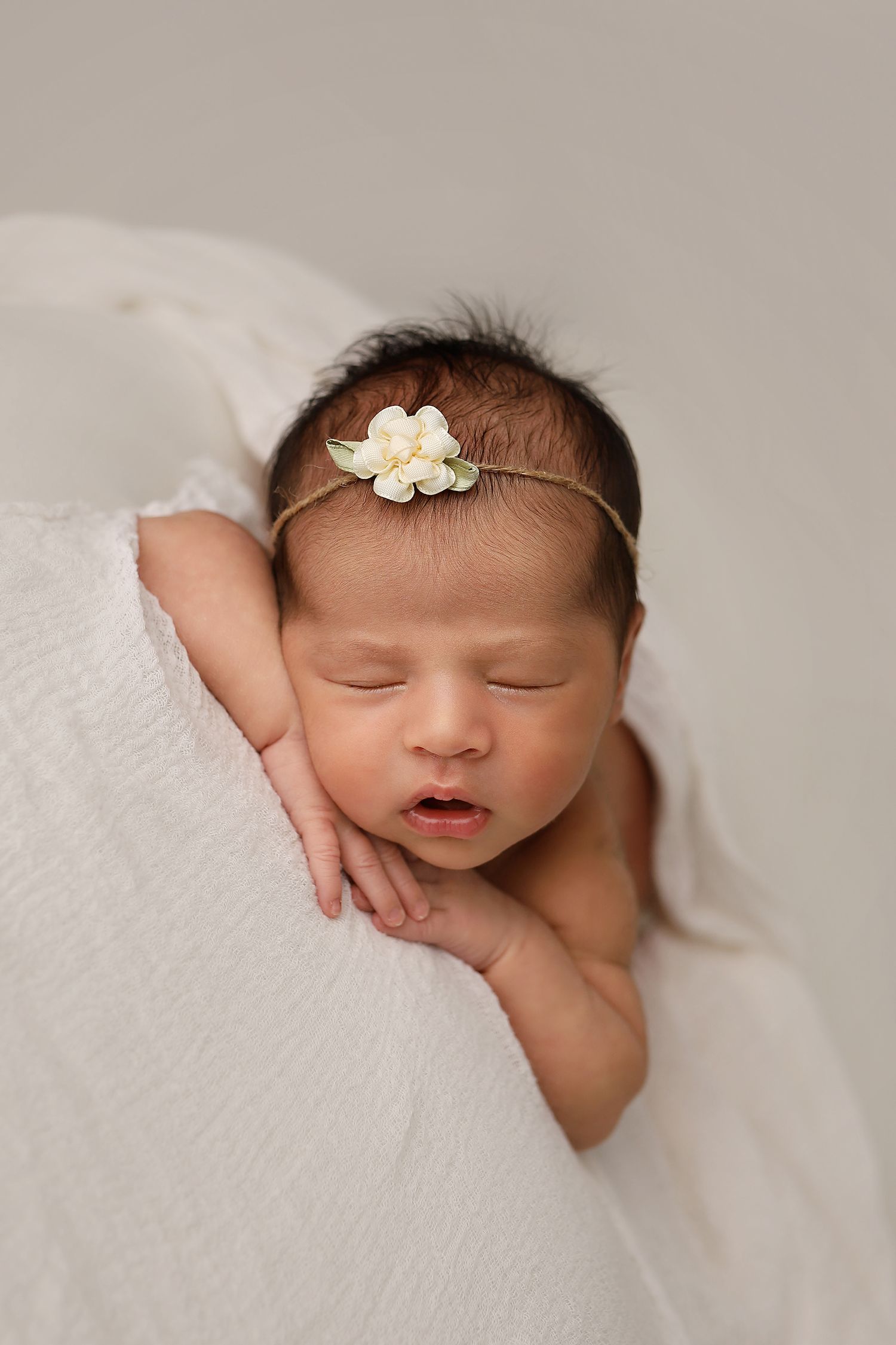 Sleeping newborn baby nestled in soft white blankets with a delicate floral headband.