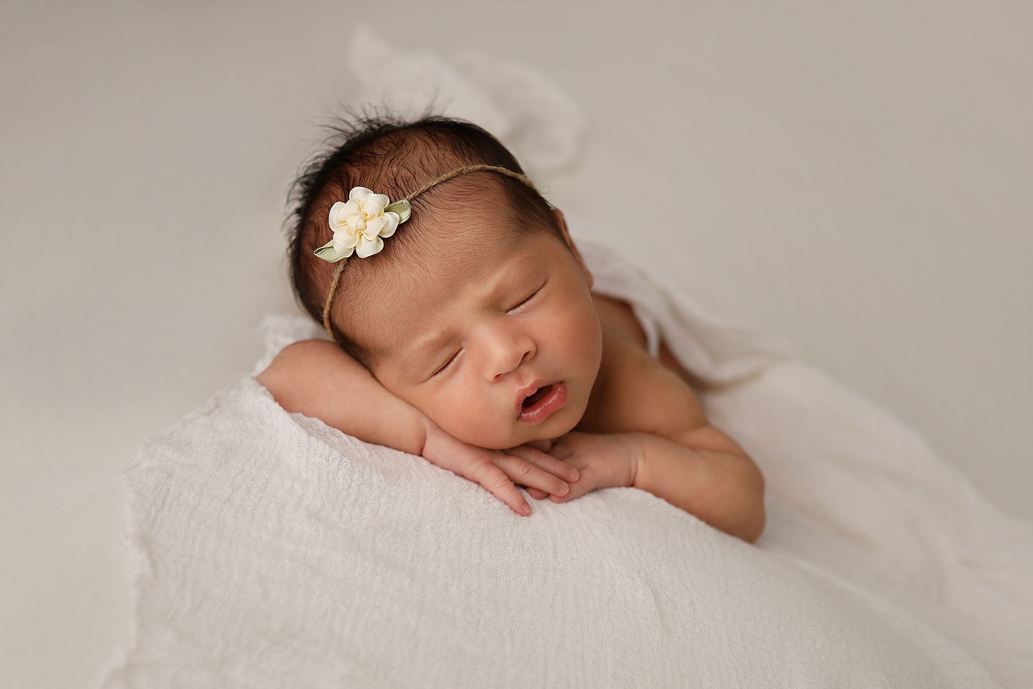 A sleeping newborn baby nestled in soft white fabric with a delicate flower hair accessory. Utah County newborn photography