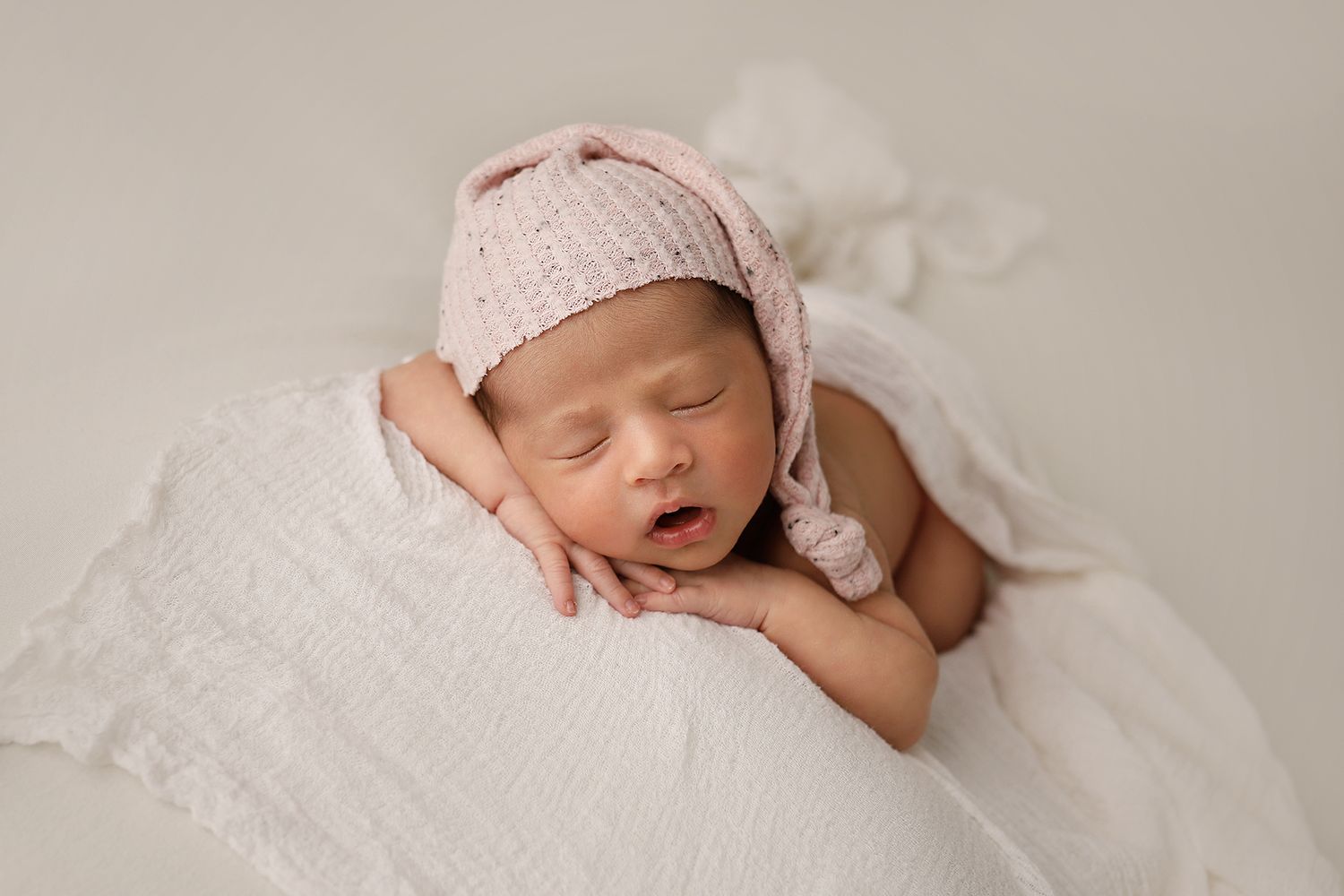 A sleeping newborn wearing a pink knit hat rests peacefully on soft white fabric.
