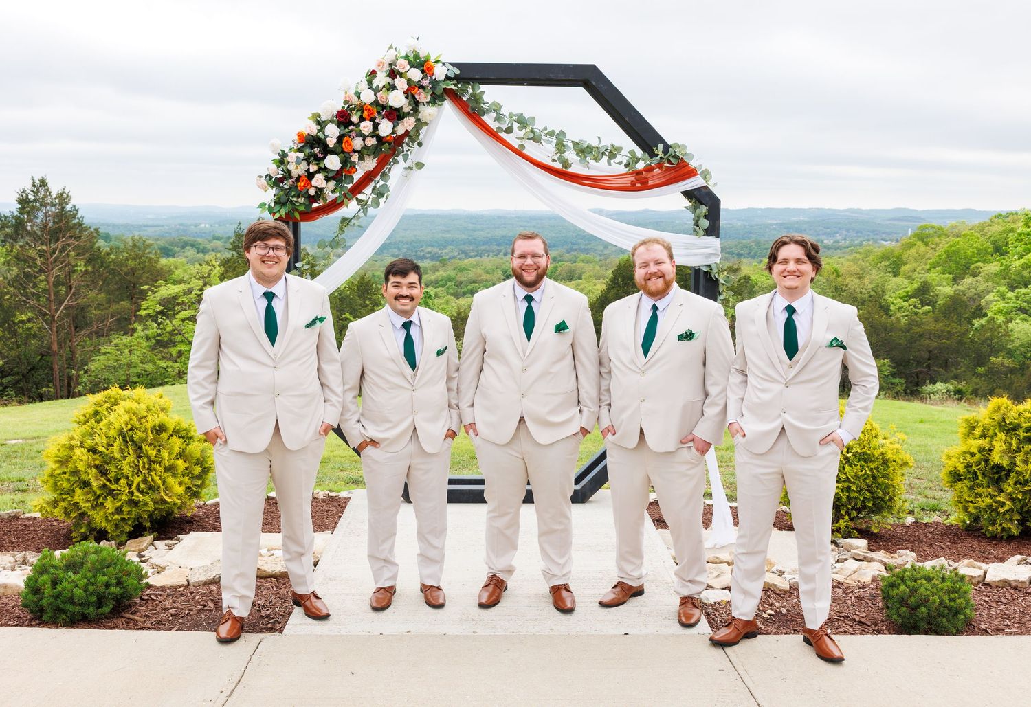 Five groomsmen in beige suits and navy blue ties stand together under a floral arch at an outdoor mountain venue.