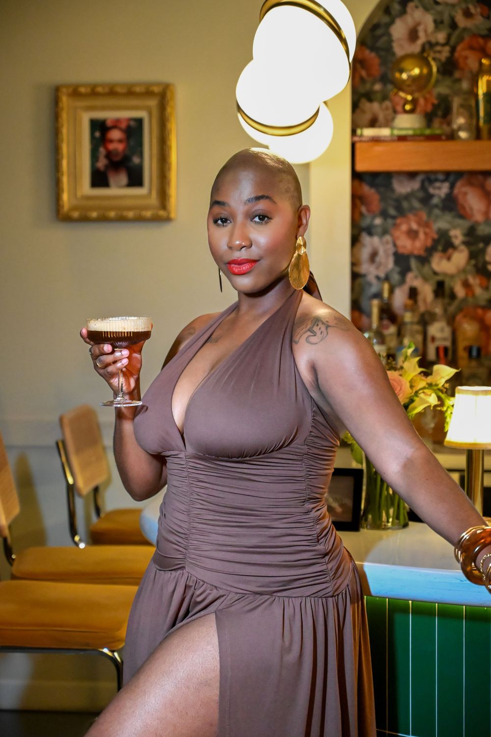 Woman in mauve halter dress poses elegantly at restaurant with cocktail and modern decor.