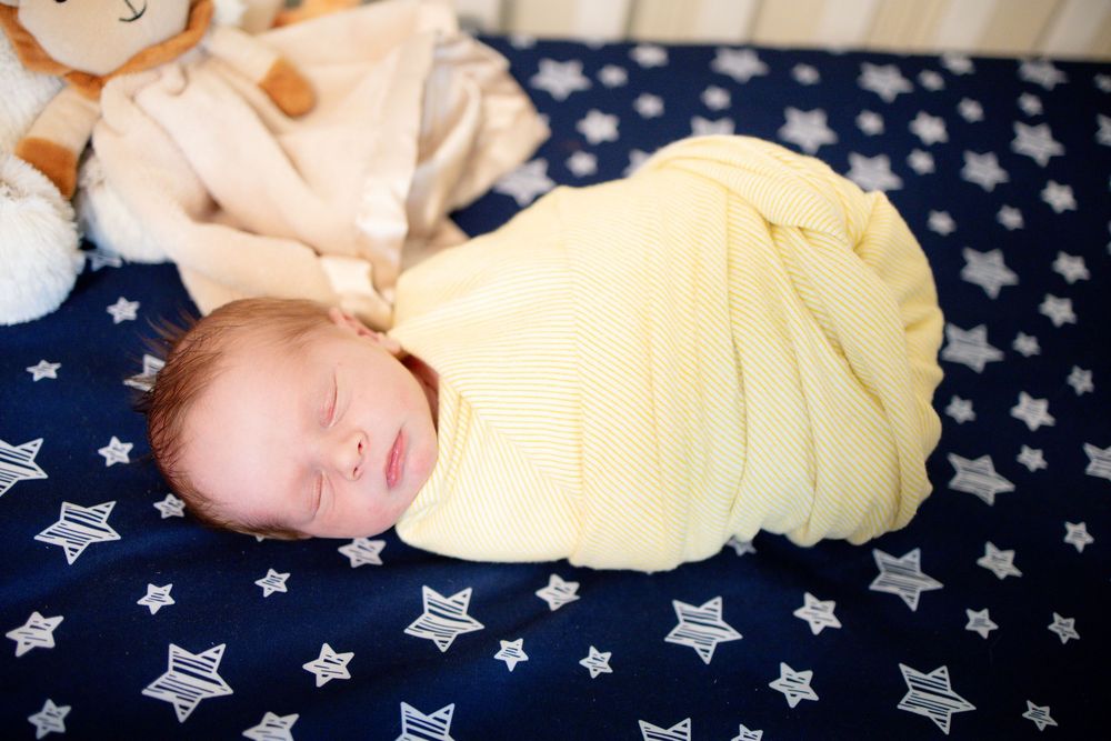 Newborn baby swaddled in yellow blanket sleeping in crib with star bedding and stuffed animal.