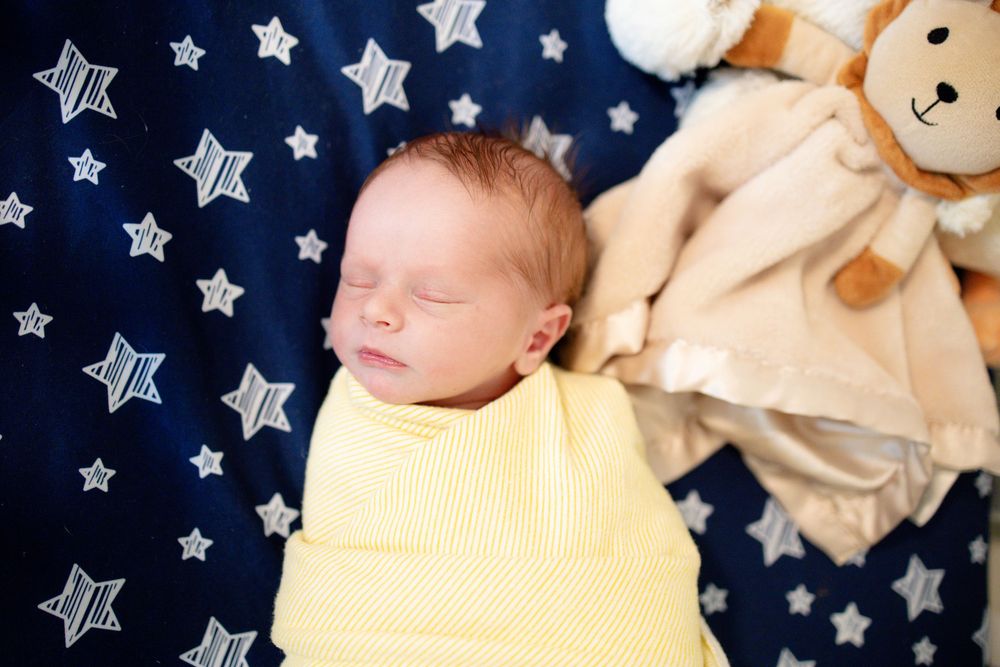 Sleeping newborn baby swaddled in yellow beside a soft teddy bear on star-print blanket.
