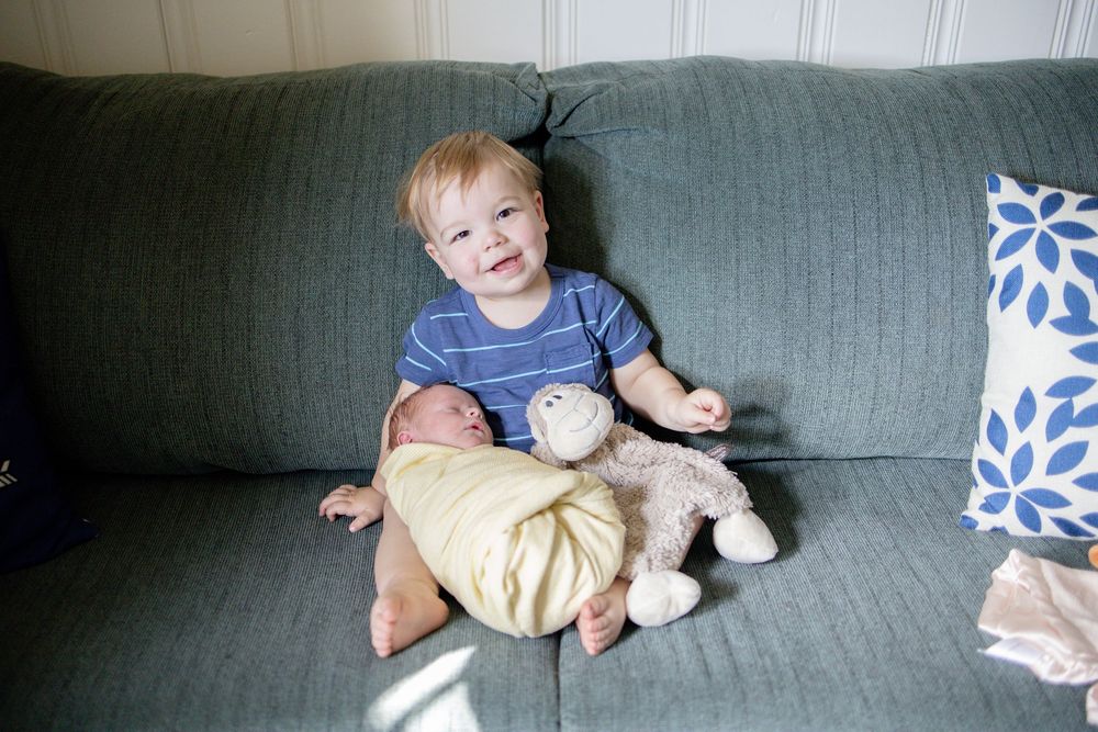 Smiling toddler boy sitting on gray sofa holding newborn sibling and stuffed animal.