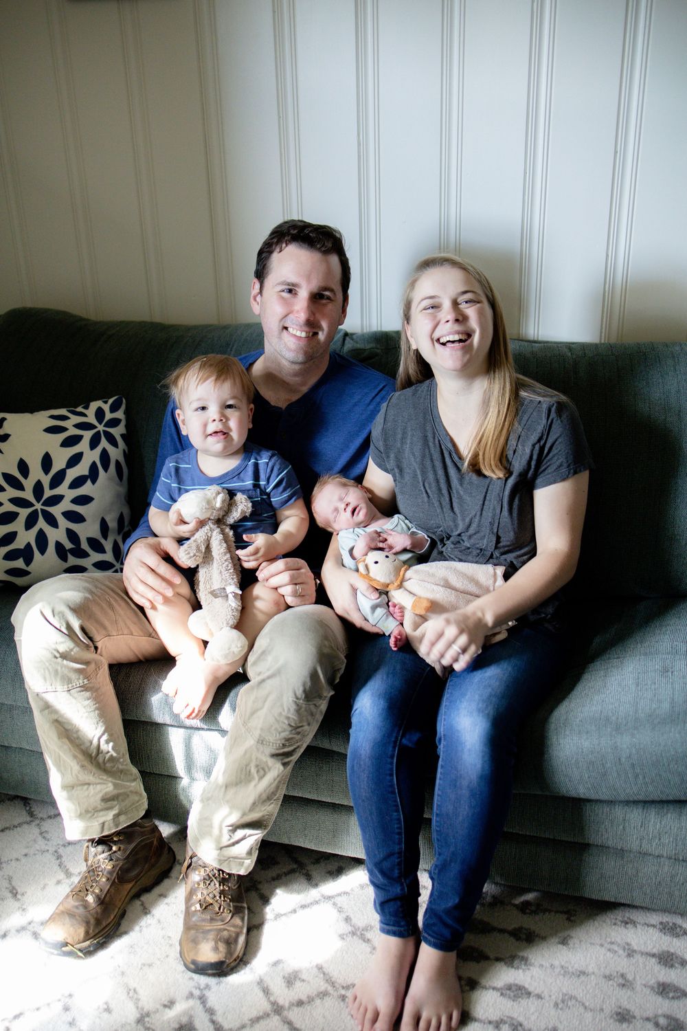 Happy young family with toddler and newborn baby smiling together on gray sofa.