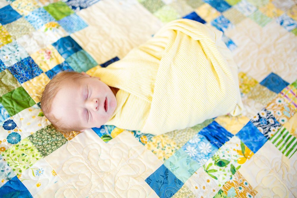 A peacefully sleeping newborn wrapped in a yellow blanket lies on a colorful patchwork quilt.