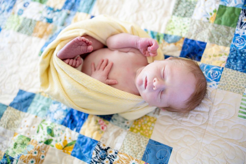 Newborn baby swaddled in yellow blanket lying on colorful patchwork quilt.