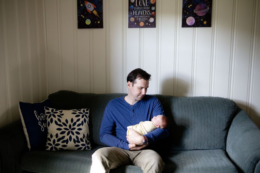 Father sitting on couch holding newborn baby wrapped in white blanket.
