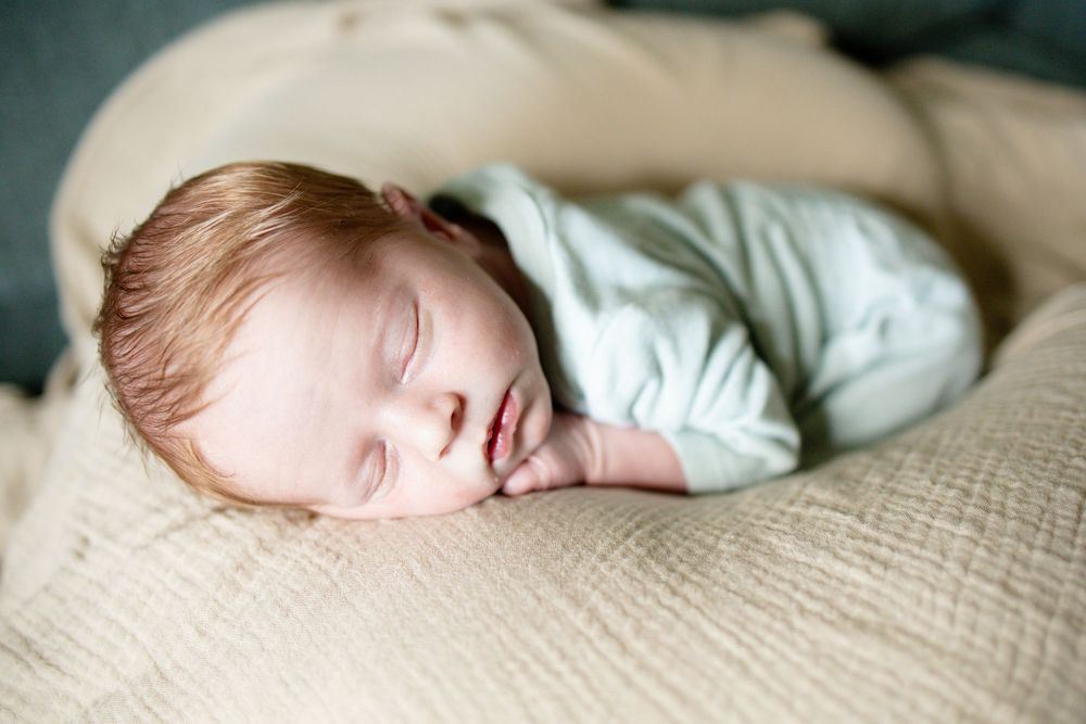 Sleeping newborn baby in mint onesie resting on beige textured blanket.
