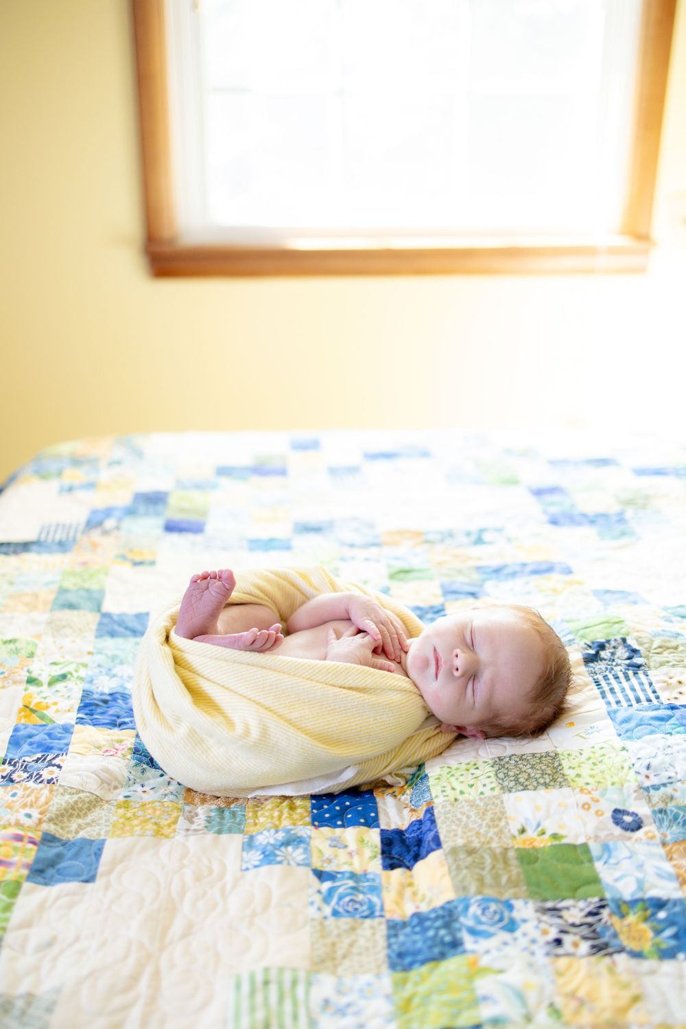 Swaddled newborn baby sleeping peacefully on colorful patchwork quilt near window.