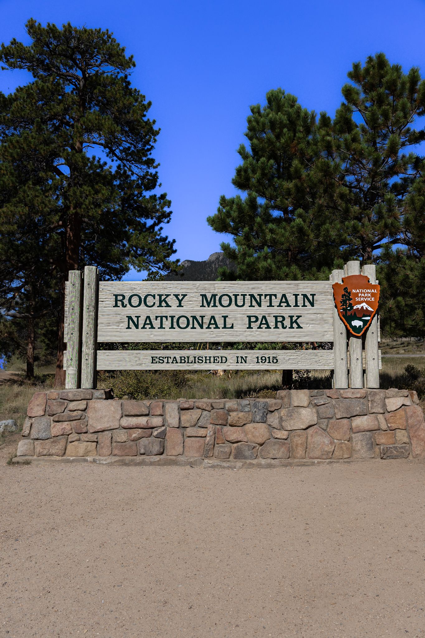 Rocky Mountain National Park entrance sign mounted on stone wall against blue sky.
