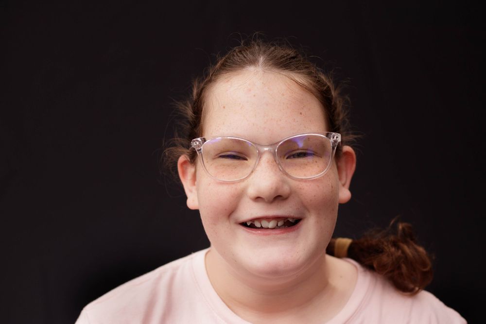 Smiling young girl with clear-framed glasses and a light pink top against a dark background.