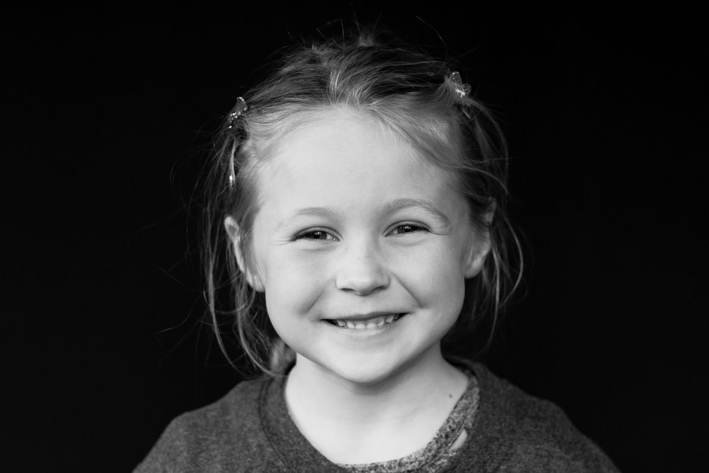 Black and white portrait of a smiling young girl with hair clips against a dark background.