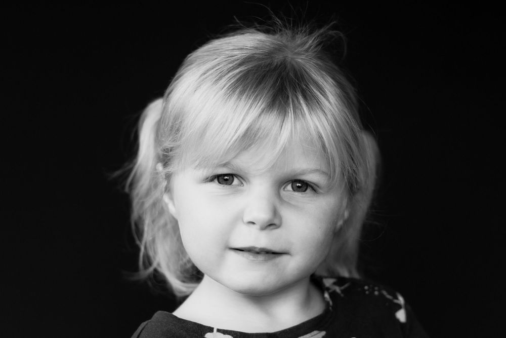 Black and white portrait of a young blonde toddler with a gentle expression on a dark background.