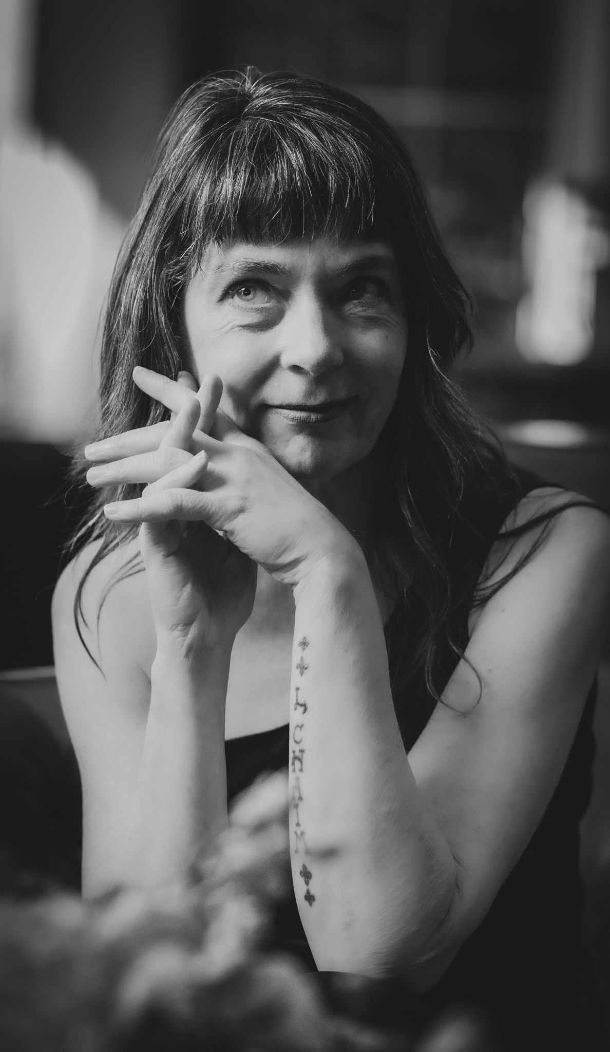 Black and white portrait of a thoughtful woman with bangs and forearm tattoo resting her chin on her hands.