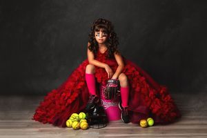 Young girl in red ruffle gown, pink knee socks, and cleats poses with softball gear in a studio portrait.