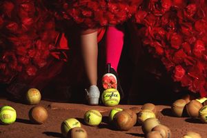 Person in pink socks and white sneakers standing among scattered softballs on dirt, surrounded by red floral backdrop.