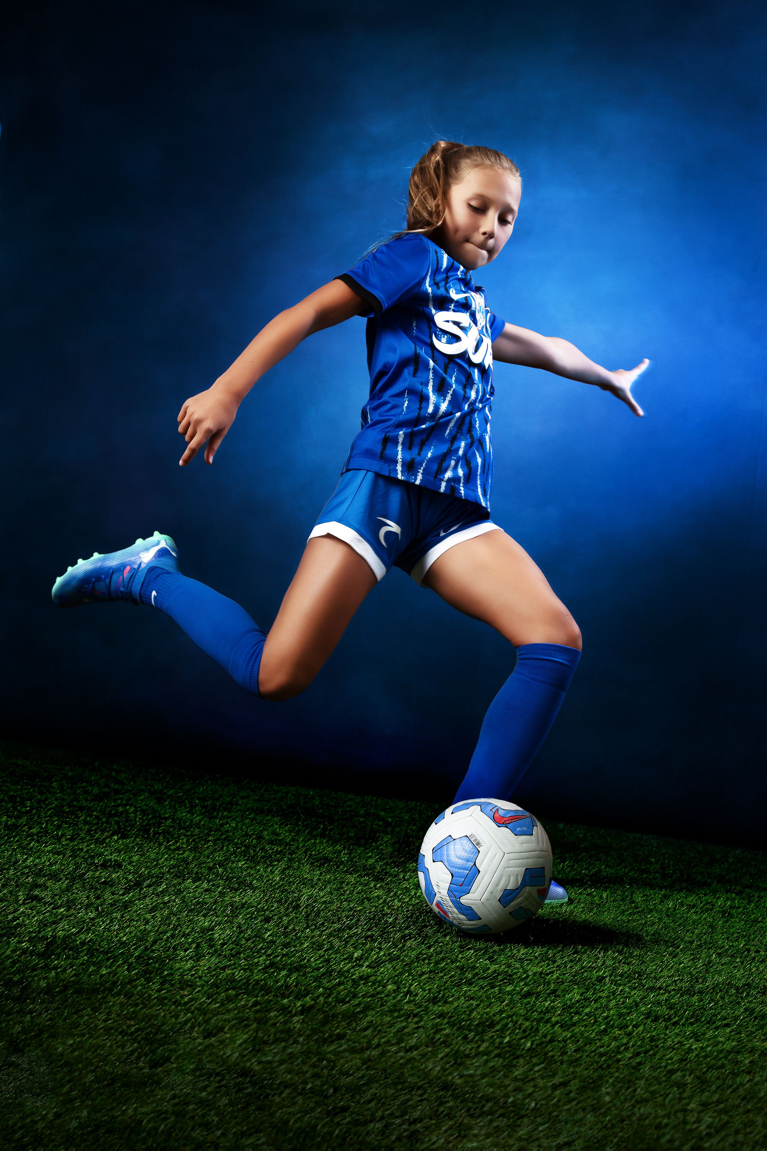 Young girl in blue soccer uniform kicking a ball on green grass field with dramatic blue background lighting.