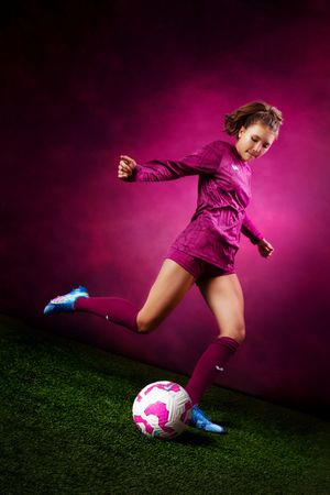 Young female soccer player in pink uniform kicking a ball on green grass against a vibrant pink background.