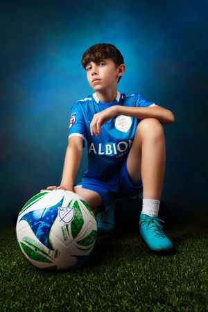 Youth soccer player in blue jersey sits with ball on artificial turf field.