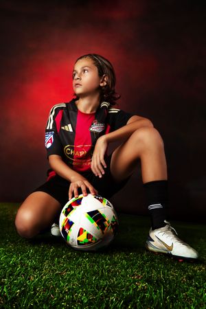 Youth soccer player in red and black jersey resting with ball on grass field with dramatic red lighting.