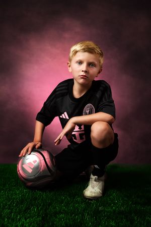 Youth athlete in black uniform poses with soccer ball on grass against pink backdrop.