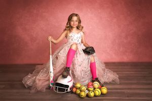 Young girl in pink gown and hockey gear poses with stick, helmet, gloves, and apples on pink background.