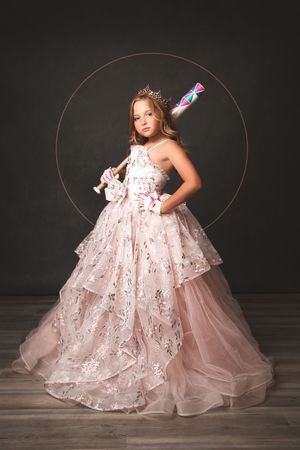 Young girl in pink floral ball gown with tiara holds doll, posing elegantly against dark studio backdrop.